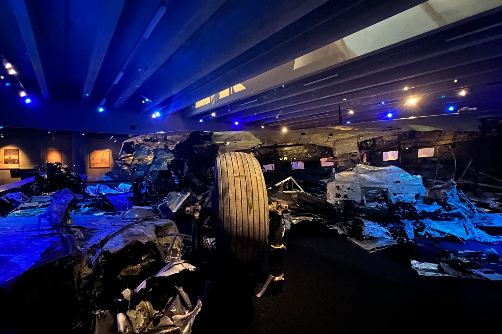 Museihall i blått ljus med vrakdelar av ett flygplan och ett stort hjul i mitten